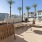 Outdoor lounge area with woven chairs, a cushioned sofa, and a round coffee table, overlooking a pool with modern townhomes in the background